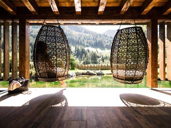 Ruhe im eigenen Takt Zwei große Hängesessel aus geflochtenem Rattan mit Blick auf einen natürlichen Badeteich und eine bewaldete Berglandschaft im Hintergrund.