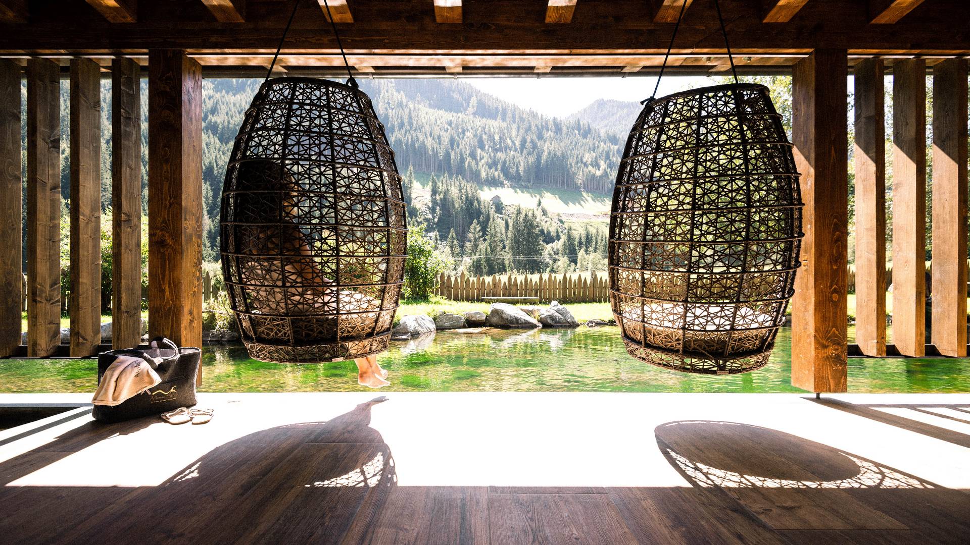 Ruhe im eigenen Takt Zwei große Hängesessel aus geflochtenem Rattan mit Blick auf einen natürlichen Badeteich und eine bewaldete Berglandschaft im Hintergrund.